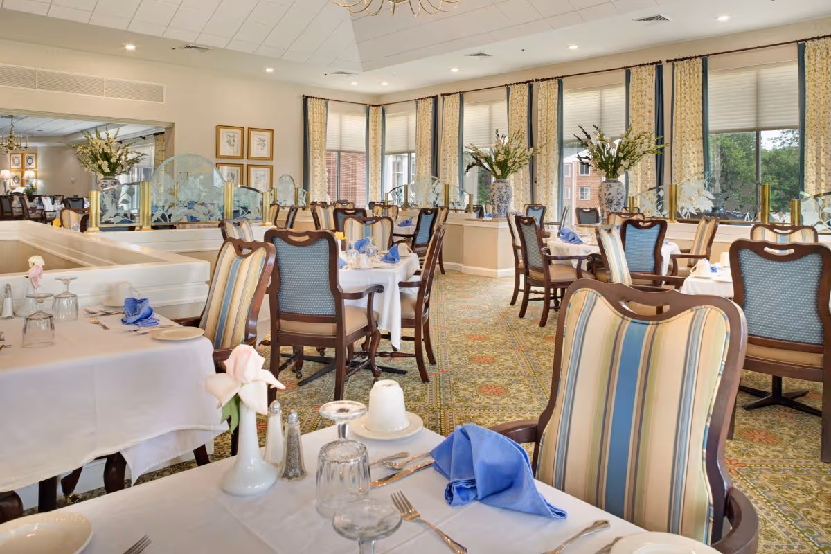 Sunlit formal dining room with arranged tables, upholstered chairs, place settings, and floral centerpieces.
