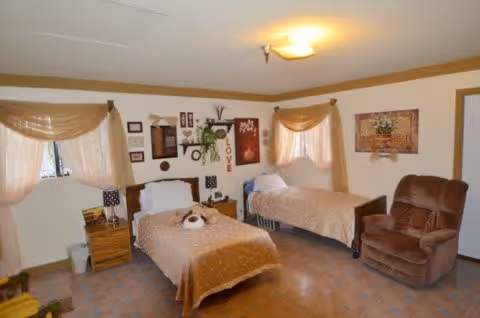A cozy bedroom with two single beds covered in beige blankets, a brown recliner chair, two windows with sheer curtains, and various framed decorations on the walls.