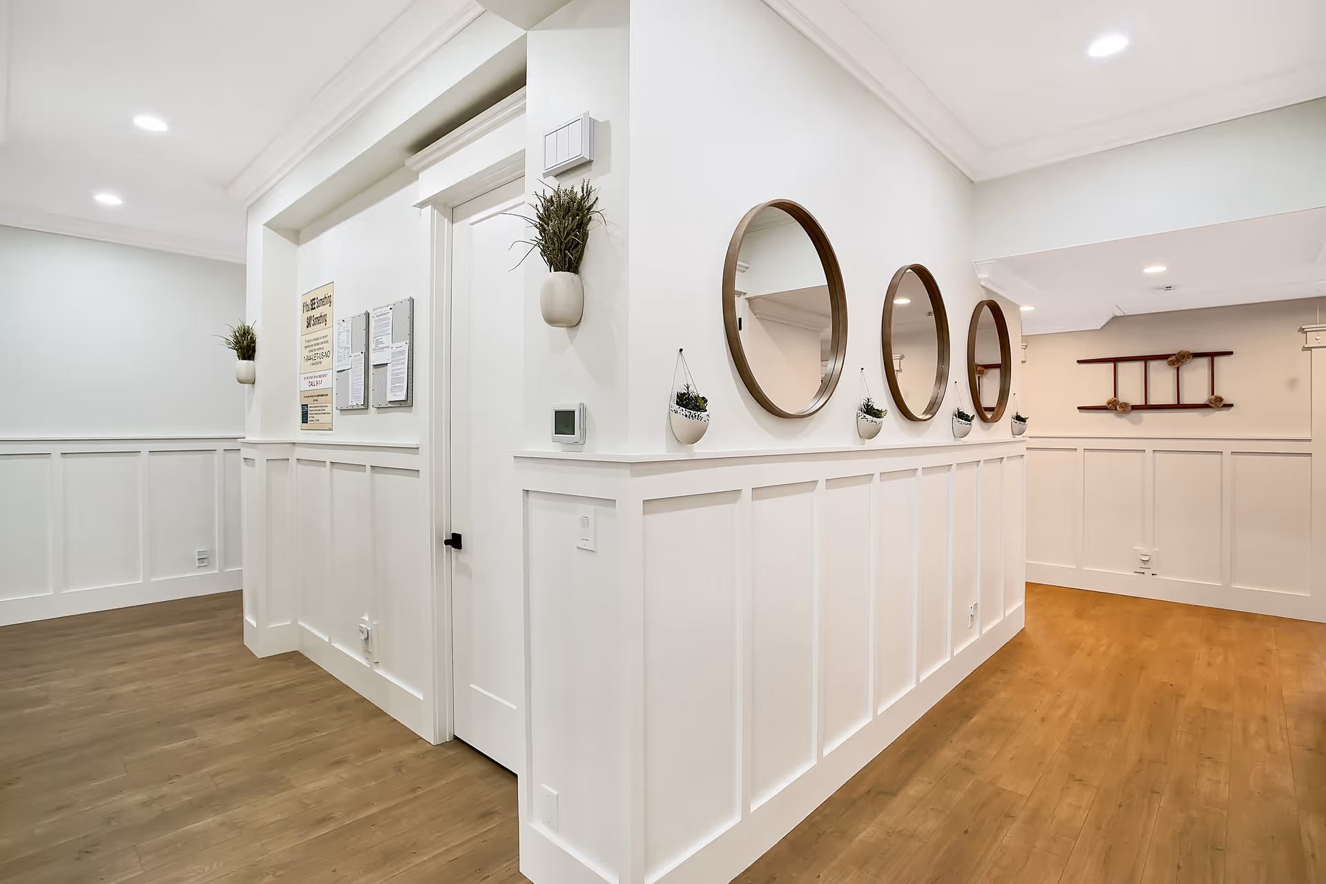 Bright interior hallway with white wainscoting, round mirrors, potted plants, and wood flooring.
