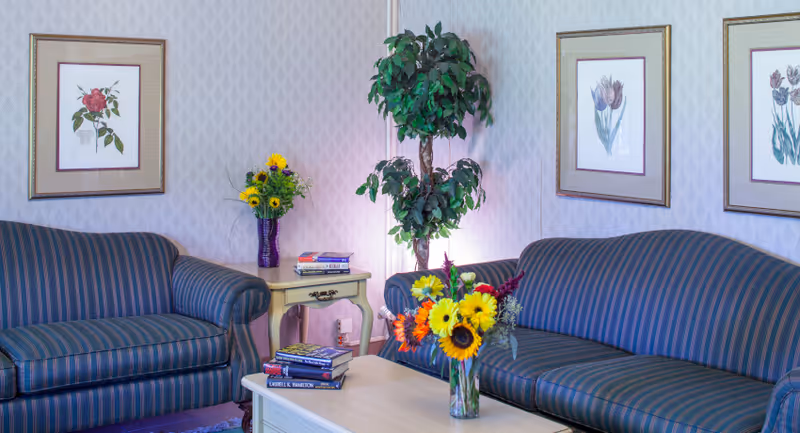 A cozy living room area with two blue striped sofas facing each other, a light-colored wooden coffee table with a vase of colorful flowers and a few books on it. There is a small side table with more books and a purple vase with yellow flowers. A tall green potted plant is placed between the sofas. The walls are decorated with framed floral artwork.