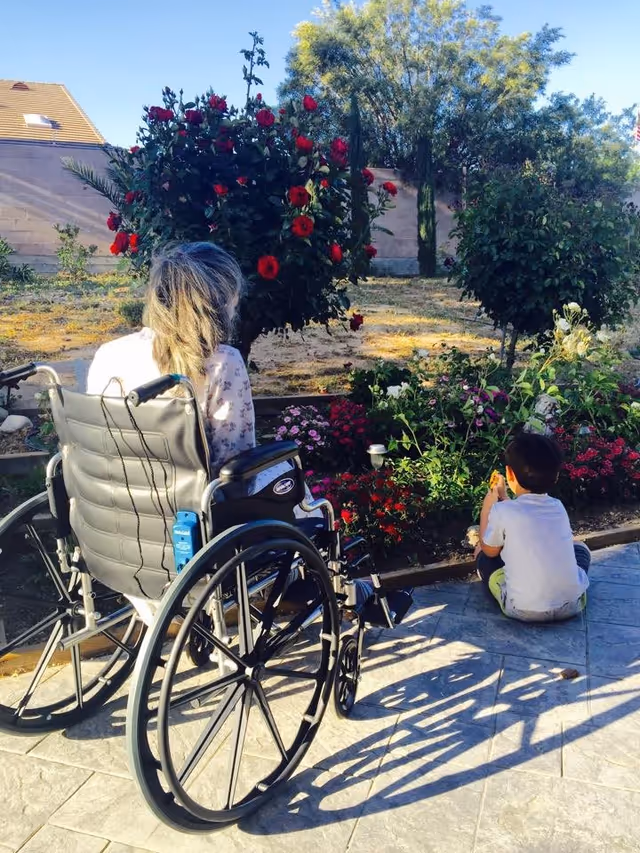 An elderly person with gray hair sitting in a wheelchair facing a garden with blooming red and pink flowers, accompanied by a young child sitting on the ground nearby, both enjoying the outdoor garden area on a sunny day.