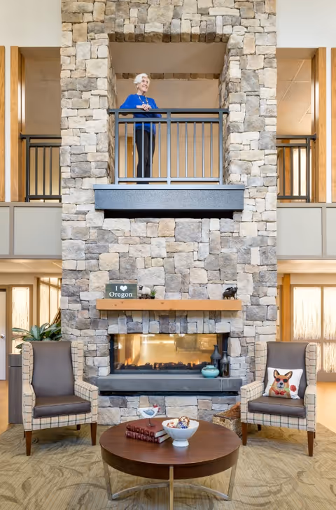 A senior woman stands on an upper balcony overlooking a cozy living room area with a stone fireplace. The fireplace is built with large gray stones and has a wooden mantel decorated with small items including a sign that says 'I ♥ Oregon'. Below the fireplace are two cushioned armchairs with patterned upholstery, one with a pillow featuring a dog illustration. A round wooden coffee table with books and a bowl sits in front of the fireplace. The room has large windows allowing natural light to fill the space.