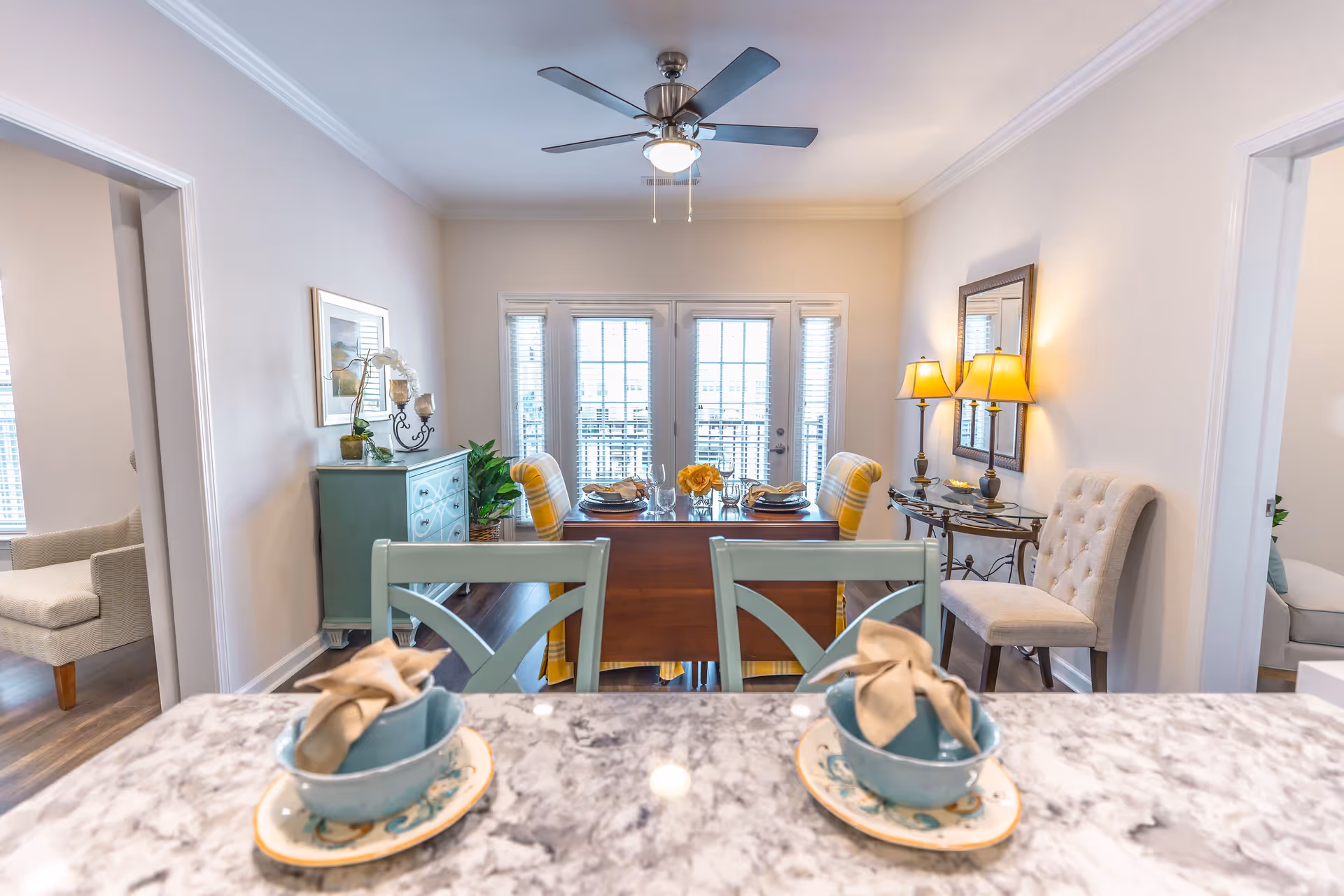 View of a bright dining room with a wooden dining table set for four, two yellow plaid chairs at the table, a ceiling fan with light above, and a sideboard with plants and decorative items. The photo is taken from a kitchen counter with two place settings in the foreground. There are doorways on either side leading to other rooms.