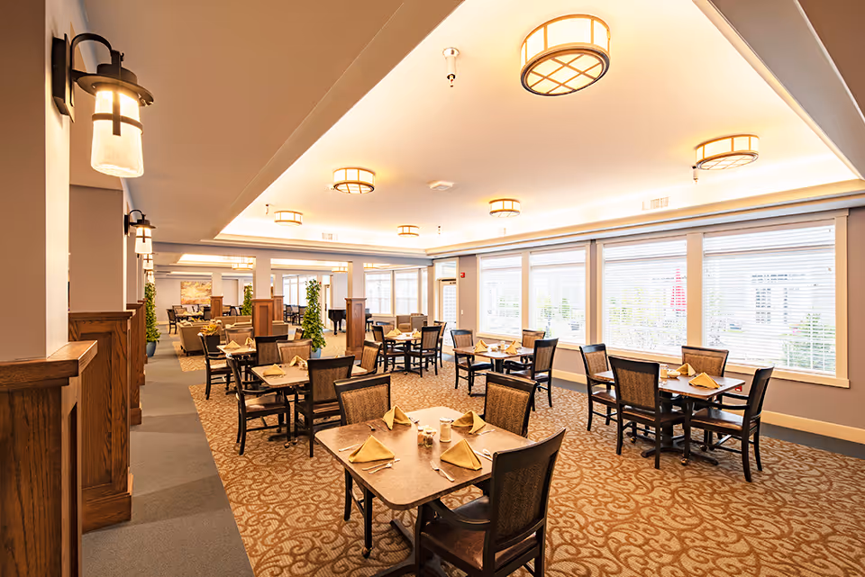 A bright and spacious dining room with multiple square tables set with folded napkins, salt and pepper shakers, and chairs. The room features large windows with blinds allowing natural light to fill the space, patterned carpet flooring, and ceiling lights. There are also wall-mounted lamps and some green plants in the background.
