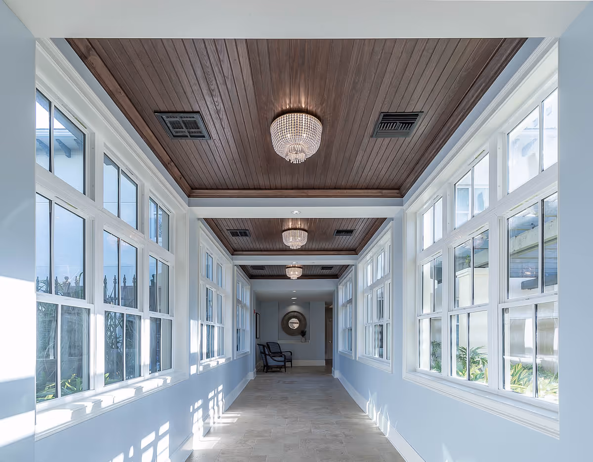 A bright hallway with large windows on both sides letting in natural light, a wooden ceiling with multiple crystal chandeliers, and a chair at the far end near a decorative mirror on the wall.