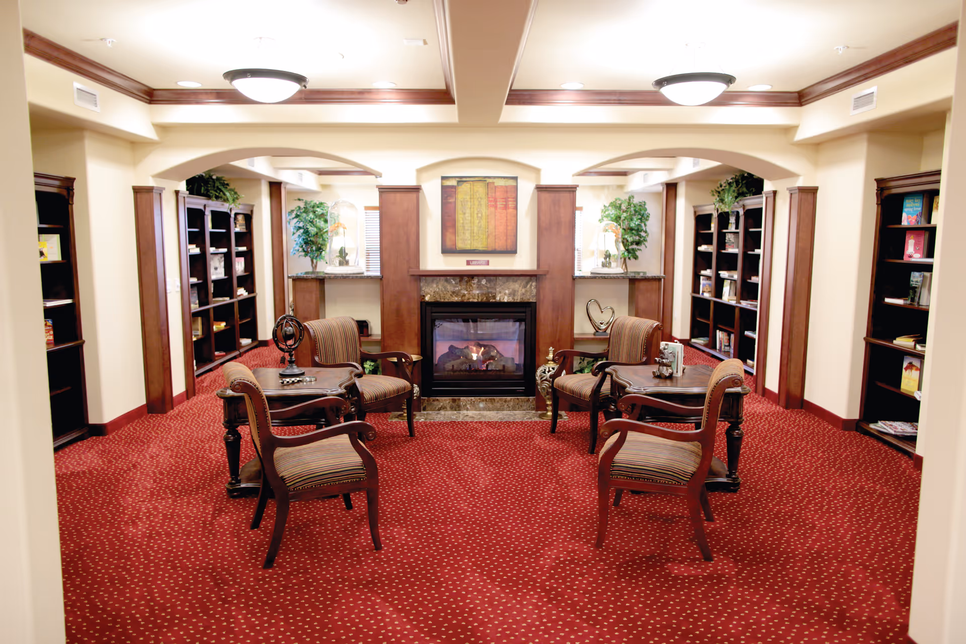 A cozy interior lounge area with a red carpet featuring small white dots, four wooden armchairs with striped cushions arranged around two small wooden tables, a fireplace with a marble surround and a framed artwork above it, and bookshelves lining the walls filled with books and decorative plants.