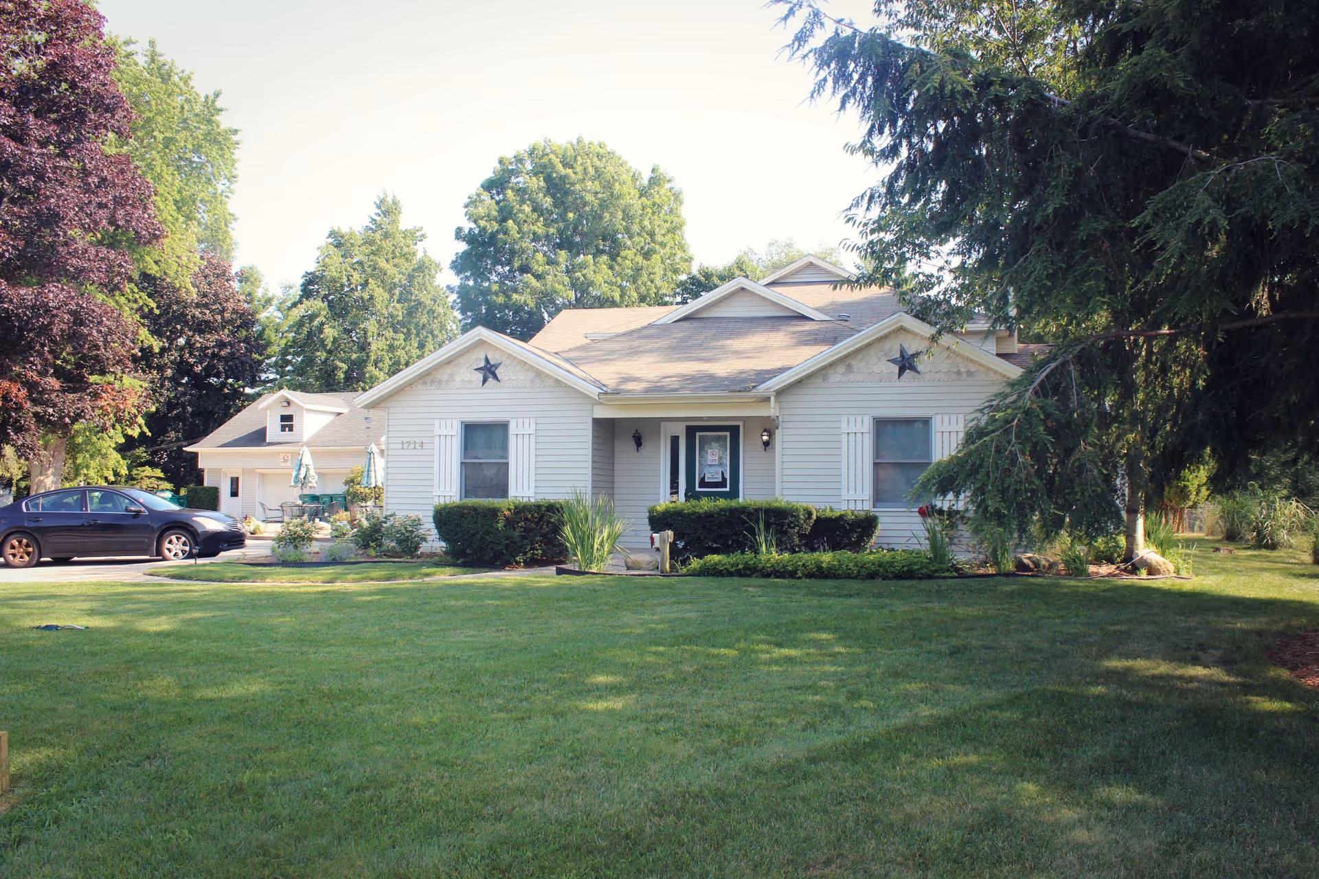 Front exterior view of a single-story residential building with light-colored siding, two windows with white shutters, a central entrance door, and decorative stars above the windows. The building is surrounded by well-maintained green lawn, bushes, and trees. A black car is parked on the driveway to the left, and there is a smaller structure behind the main building.