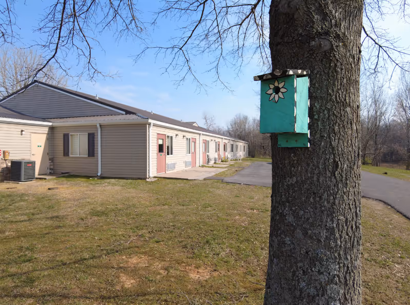 A single-story beige building with multiple doors and windows, situated on a grassy area with a paved path. In the foreground, a tree with a turquoise birdhouse decorated with a white flower is visible. The sky is clear and blue.