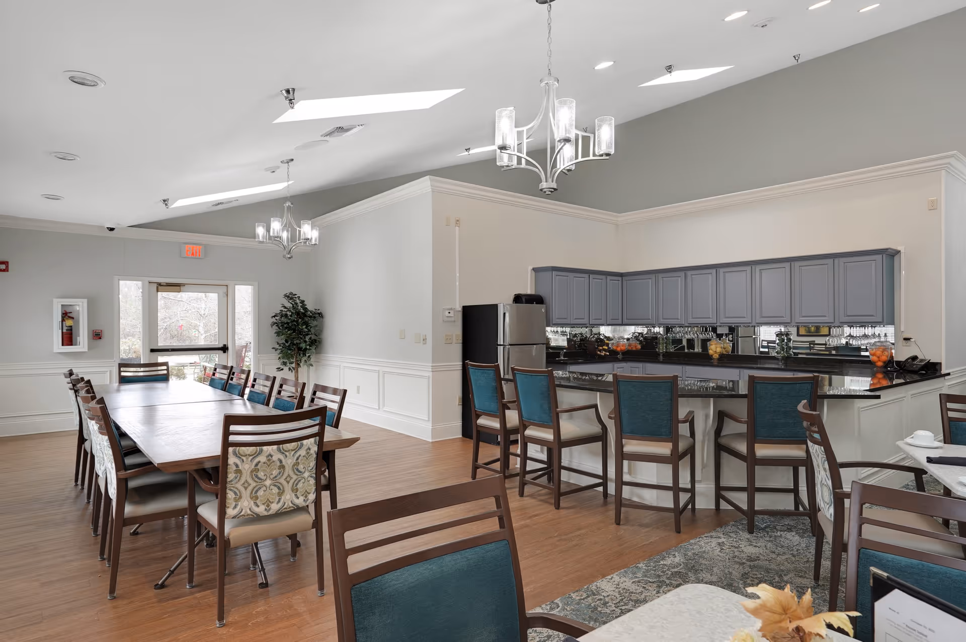 A spacious dining area with a long wooden table surrounded by chairs on the left and a kitchen counter with bar stools on the right. The kitchen has gray cabinets, a stainless steel refrigerator, and a mirrored backsplash. The room features hardwood floors, two modern chandeliers, and a door leading outside with windows on either side. A potted plant is placed near the door.