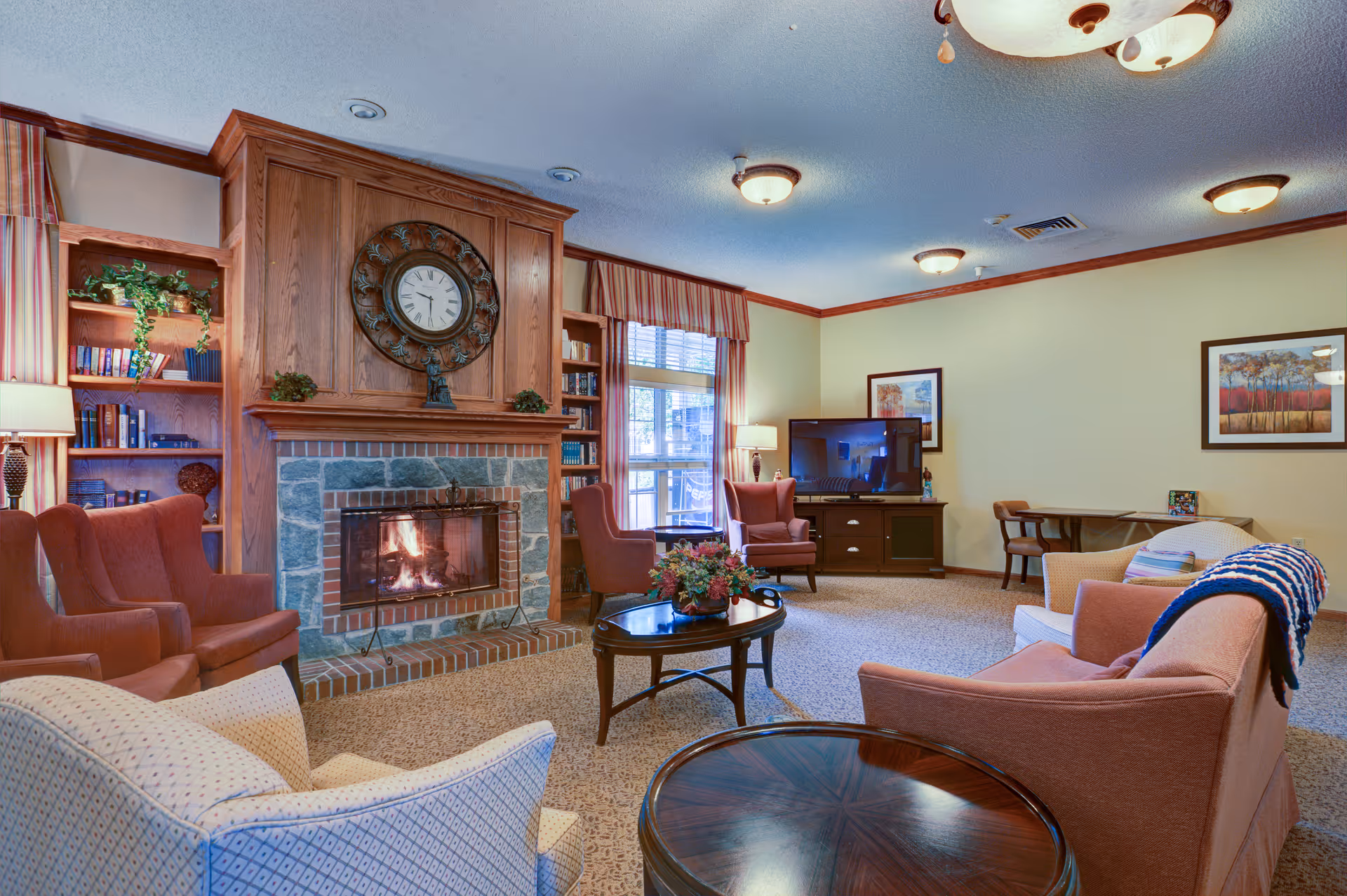 A cozy living room with a stone and wood fireplace, a large decorative clock above it, and several upholstered armchairs arranged around two wooden coffee tables. There is a TV on a wooden stand against the far wall, two framed landscape paintings, a window with striped curtains, and a small desk with a chair in the corner.
