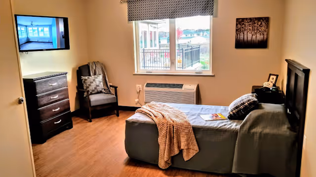 A cozy bedroom with a single bed covered with a gray bedspread and a beige throw blanket. There is a dark wooden headboard, a nightstand with a photo frame and a clock, a cushioned armchair with a patterned pillow, and a chest of drawers with a TV mounted above it. A window with a patterned valance lets in natural light and shows an outdoor view.