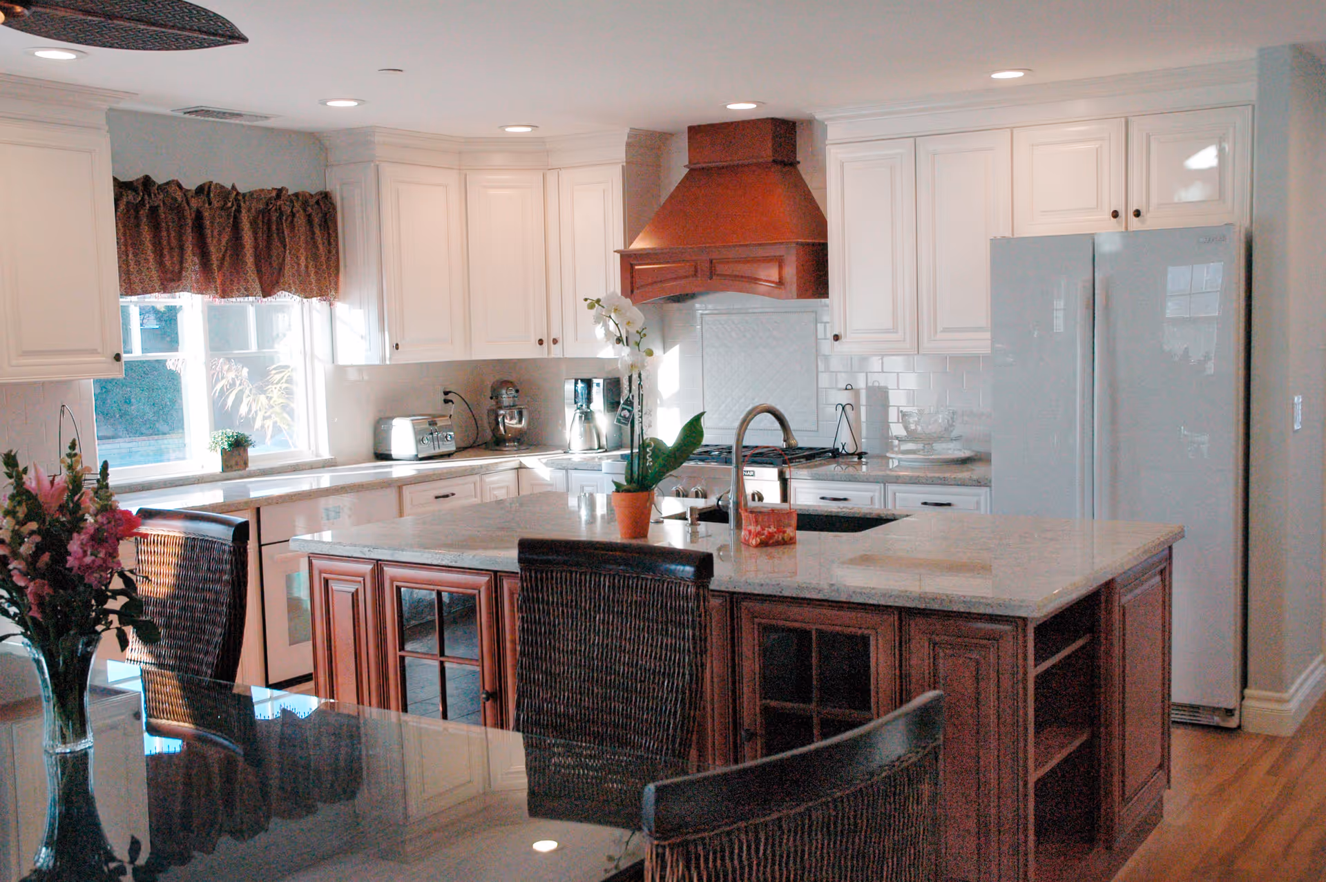 A bright kitchen with white cabinets, a large island with a marble countertop, and a wooden range hood. There are wicker chairs around the island and a glass dining table with a vase of flowers. Appliances include a refrigerator, toaster, blender, and stand mixer. A window with a brown valance lets in natural light.