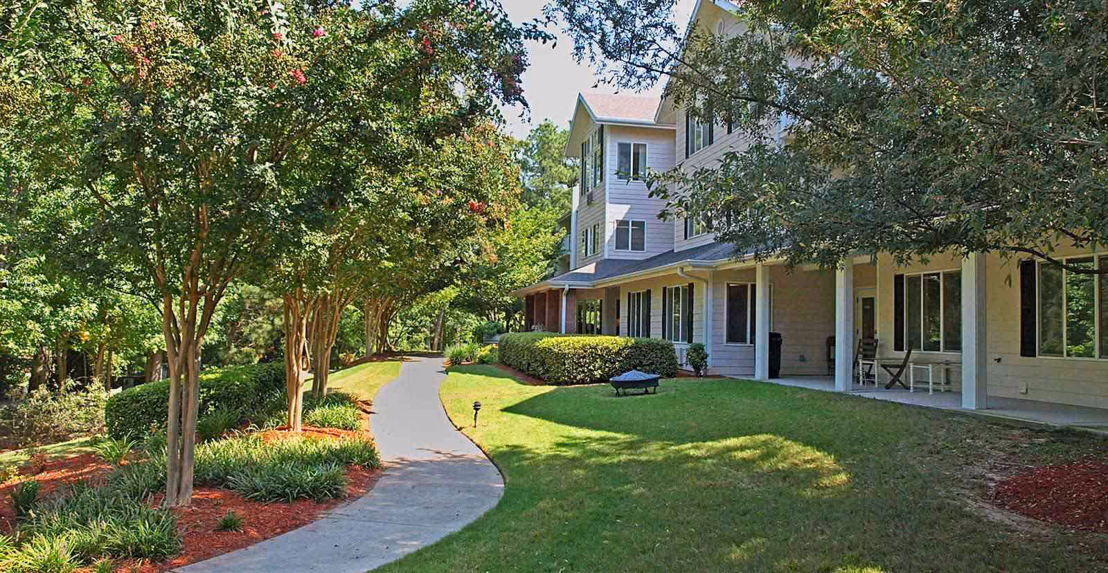 A paved walkway curves through a landscaped garden with green grass, bushes, and trees with some pink flowers. To the right is a multi-story building with a covered porch featuring chairs and tables.