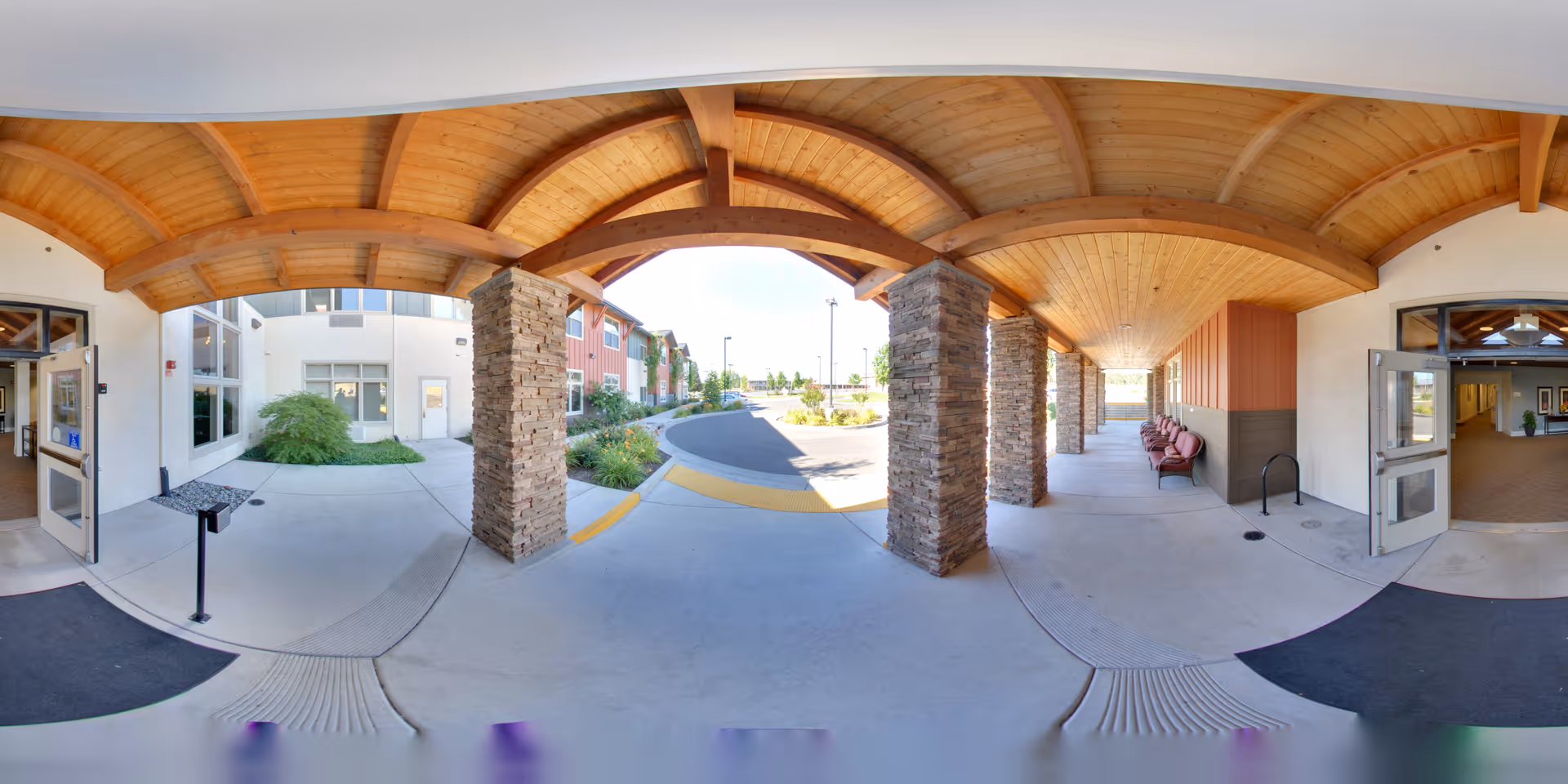 Covered entrance canopy with a curved wooden ceiling and stone pillars leading to the building's double doors and driveway, with seating along one wall.