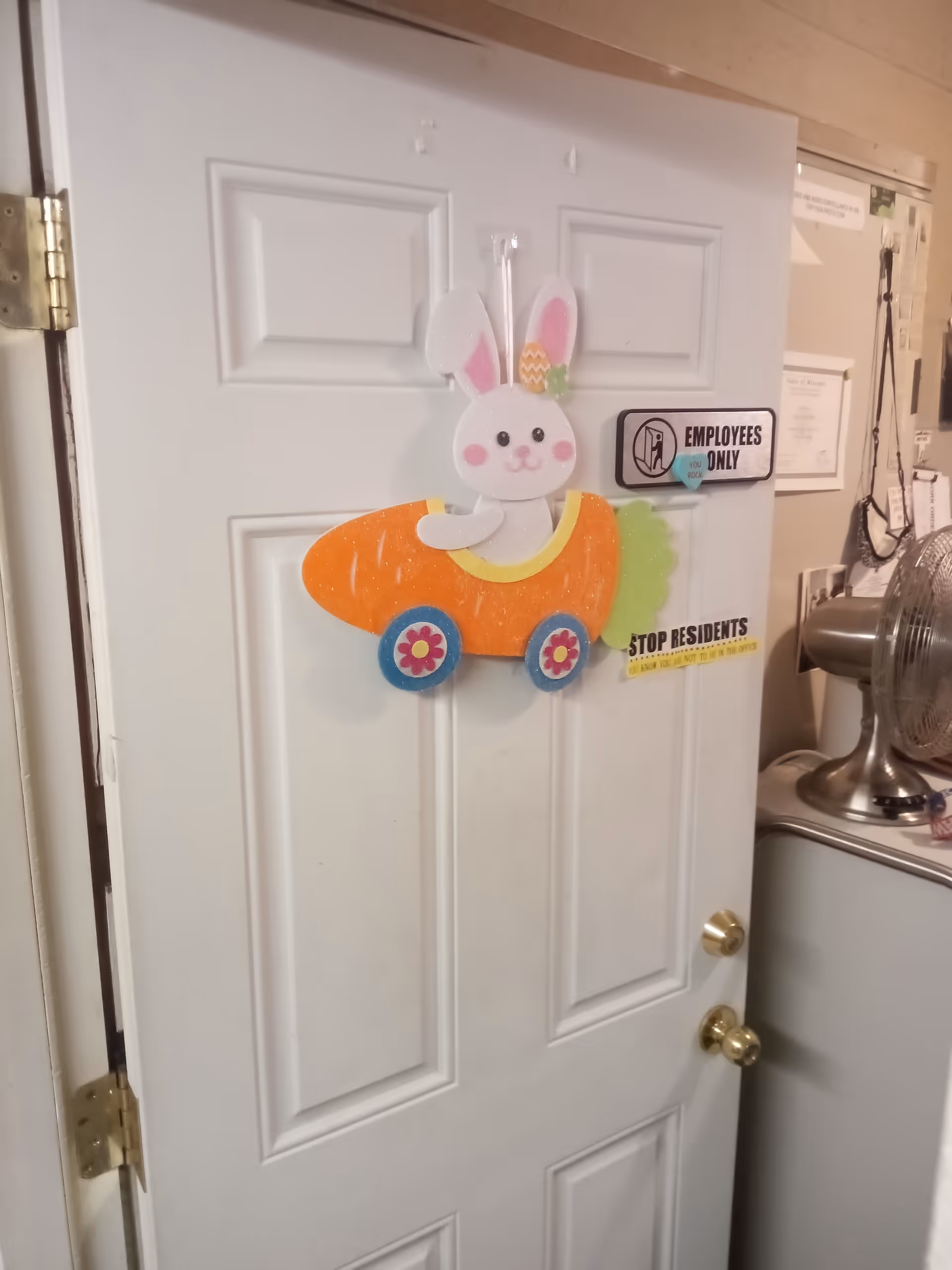 A white door with two gold-colored knobs and a hinge on the left side. The door has a decoration of a white bunny with pink cheeks and ears, sitting in an orange carrot-shaped car with blue wheels. There are two signs on the door: one says 'EMPLOYEES ONLY' with a no entry symbol, and another partially visible yellow sign that says 'STOP RESIDENTS'. To the right of the door, there is a metallic fan on top of a white appliance, and some papers and items pinned on a beige wall.
