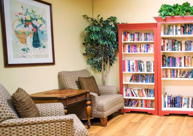 A cozy reading nook with two patterned armchairs, a wooden side table between them, a tall artificial plant in the corner, and two red bookshelves filled with books against a beige wall. A framed floral painting hangs on the wall above one of the chairs.