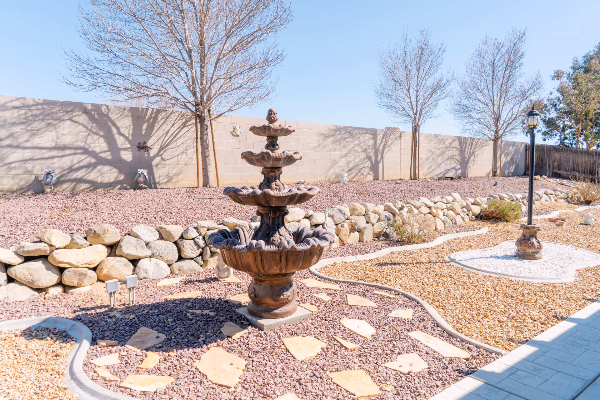 A landscaped outdoor garden area with a multi-tiered stone fountain in the center, surrounded by decorative rocks and stepping stones. There are leafless trees and a stone wall in the background, along with a vintage-style lamp post on the right side.