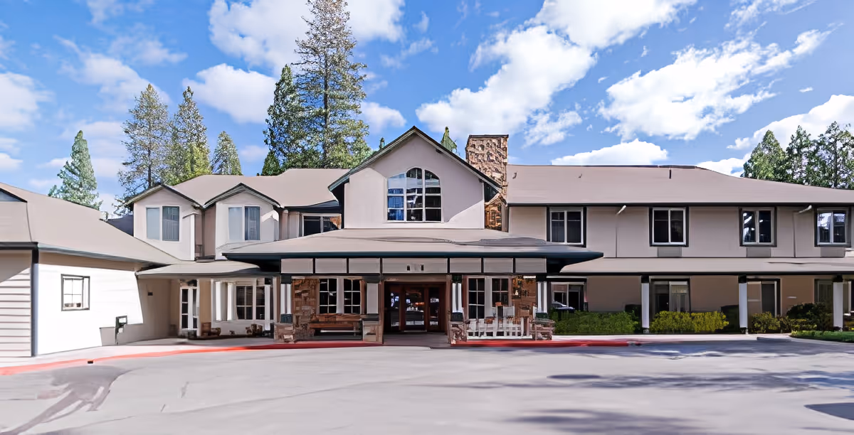 Front exterior view of Cascades of Grass Valley facility showing a two-story building with a covered entrance, large windows, stone chimney, and surrounded by tall trees under a blue sky with scattered clouds.