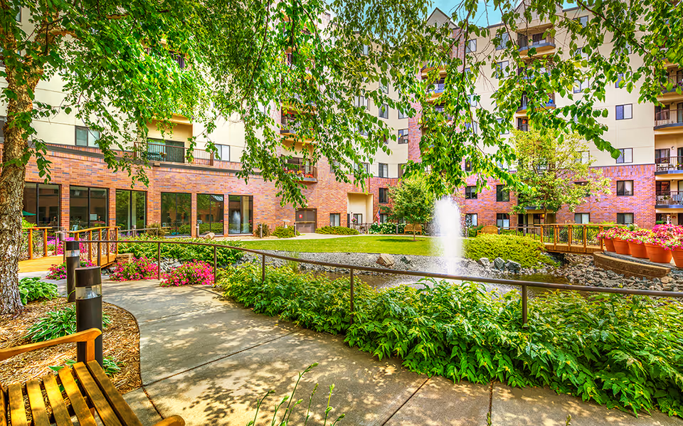 Outdoor courtyard area at Covenant Living of Golden Valley featuring a walking path, wooden bench, lush green plants, colorful flowers, a small pond with a water fountain, and a multi-story brick and beige building in the background with balconies and windows.