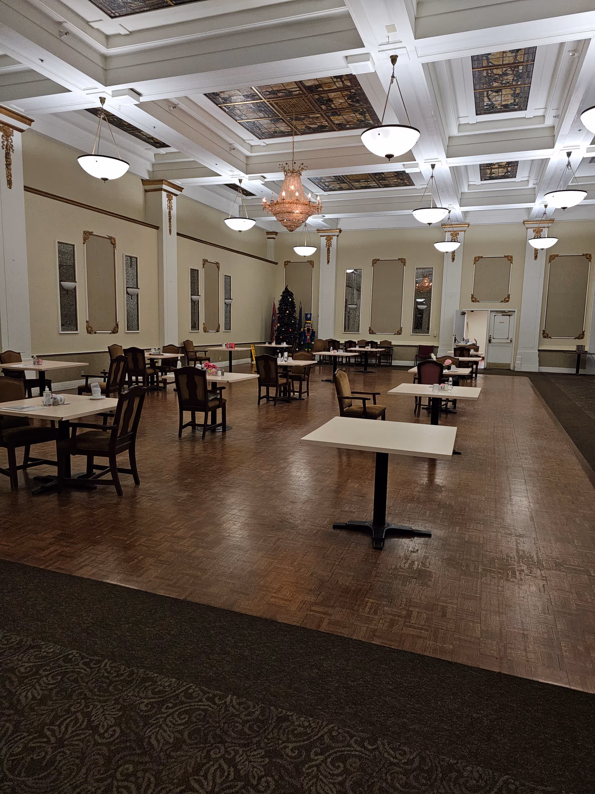A spacious dining room with multiple tables and chairs arranged neatly. The room features a high ceiling with decorative panels and several hanging light fixtures, including a large chandelier in the center. There is a Christmas tree and a nutcracker decoration in the far corner, and the walls have ornate moldings and mirrors.