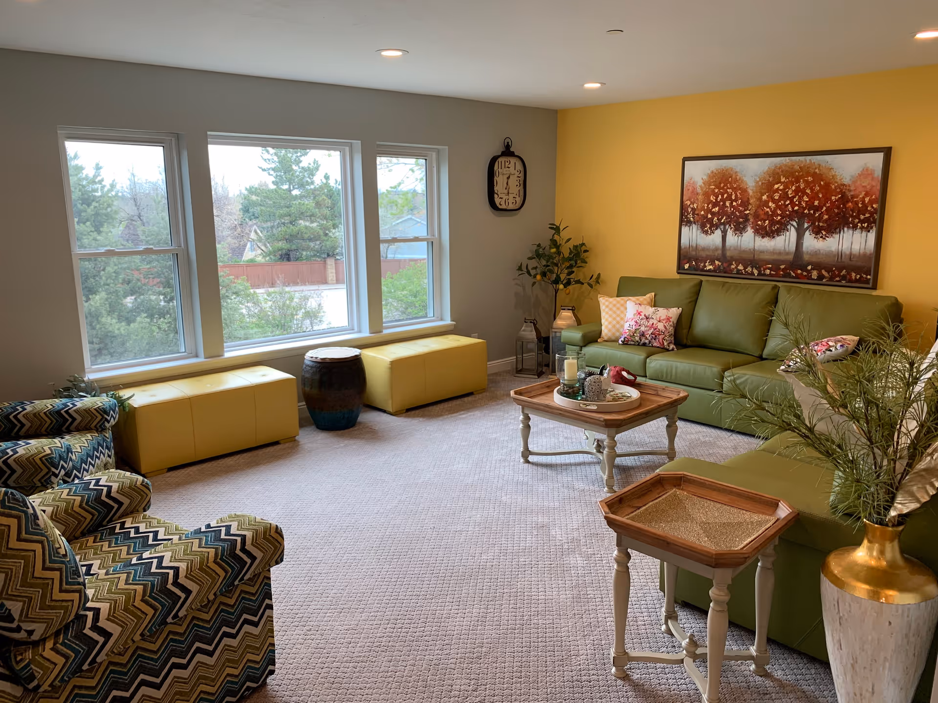 A cozy living room with a green leather sofa adorned with floral pillows, a wooden coffee table with decorative items, a patterned armchair, and two yellow ottomans by three large windows showing trees outside. The walls are painted gray and yellow, with a large painting of autumn trees on the yellow wall and a clock on the gray wall. There are also plants and lanterns adding to the decor.