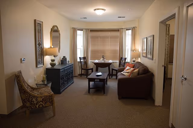 A cozy living room area in a senior living facility with a brown sofa adorned with pillows, a wooden coffee table, two wooden chairs around a small dining table near a window with blinds, a patterned armchair, a black sideboard with decorative items, a wall mirror, and warm lighting from table lamps.