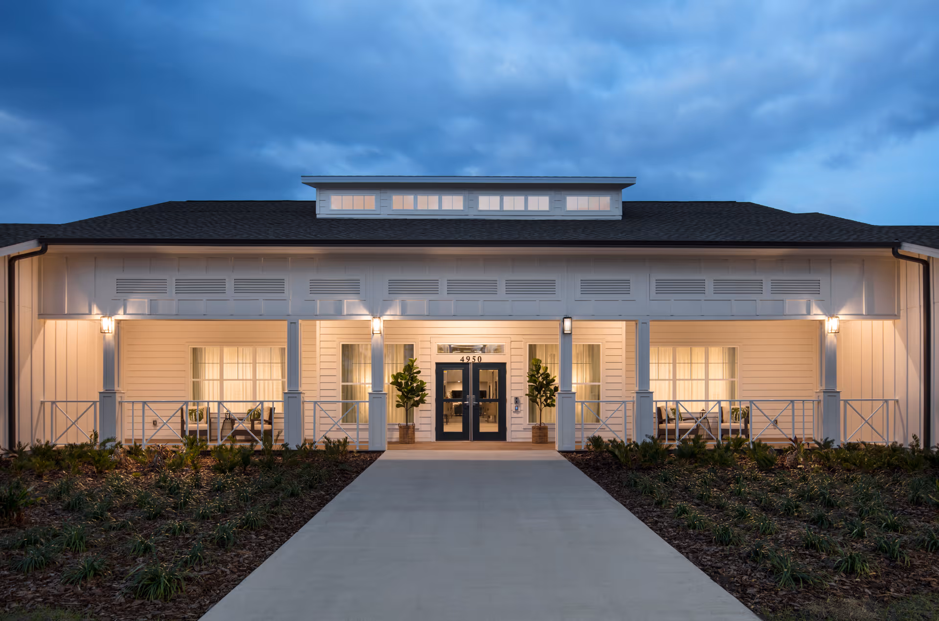 Front exterior view of a single-story senior living facility building at dusk with warm interior lights visible through windows, a central entrance with double glass doors, potted plants on either side, and a concrete walkway leading up to the entrance.