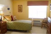 A small, neatly arranged bedroom in a nursing and rehab center with a single bed covered in a green quilt, a bedside table with a lamp, a framed picture on the wall, a window with a brown valance, an air conditioning unit below the window, and a wooden chair with a cushion.