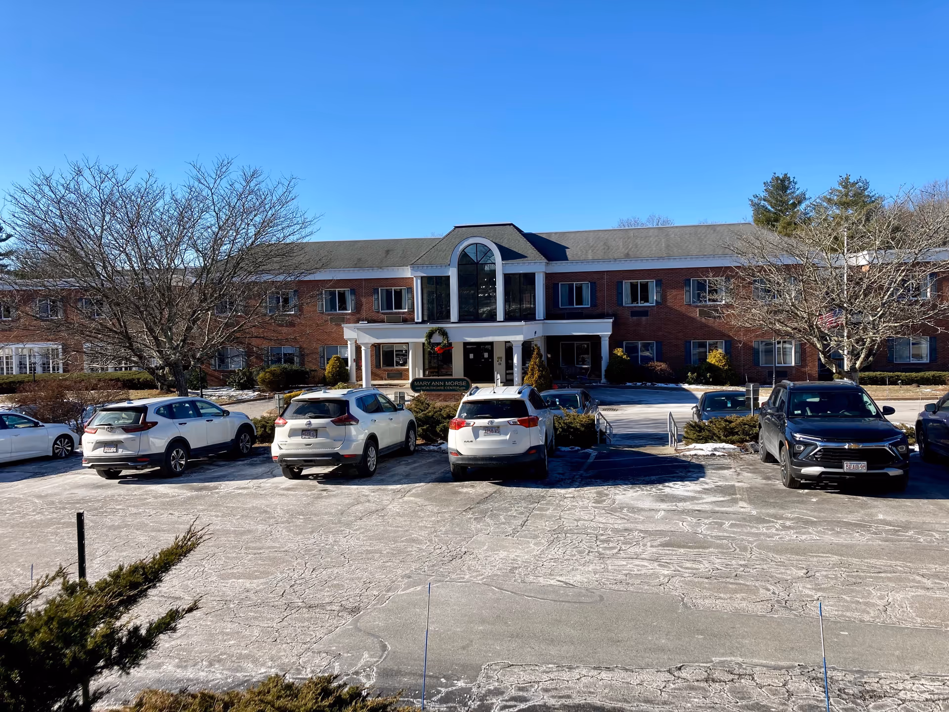 Front exterior view of Mary Ann Morse at Heritage, a two-story brick building with a central entrance featuring large windows and a covered porch. Several cars are parked in the parking lot in front of the building, and leafless trees are visible on either side under a clear blue sky.