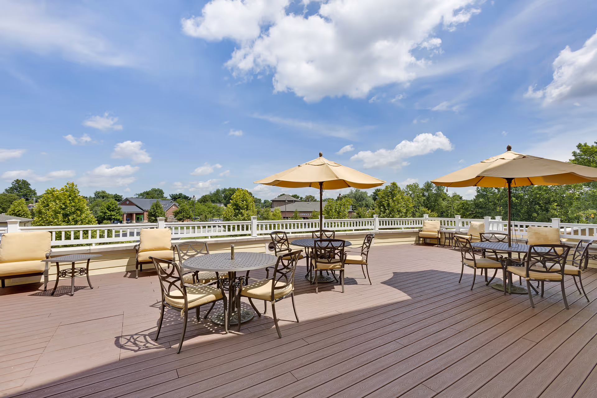 Outdoor patio area with several round tables and chairs, each table shaded by a large beige umbrella. Cushioned seating lines the perimeter against a white railing, with trees and houses visible in the background under a partly cloudy blue sky.