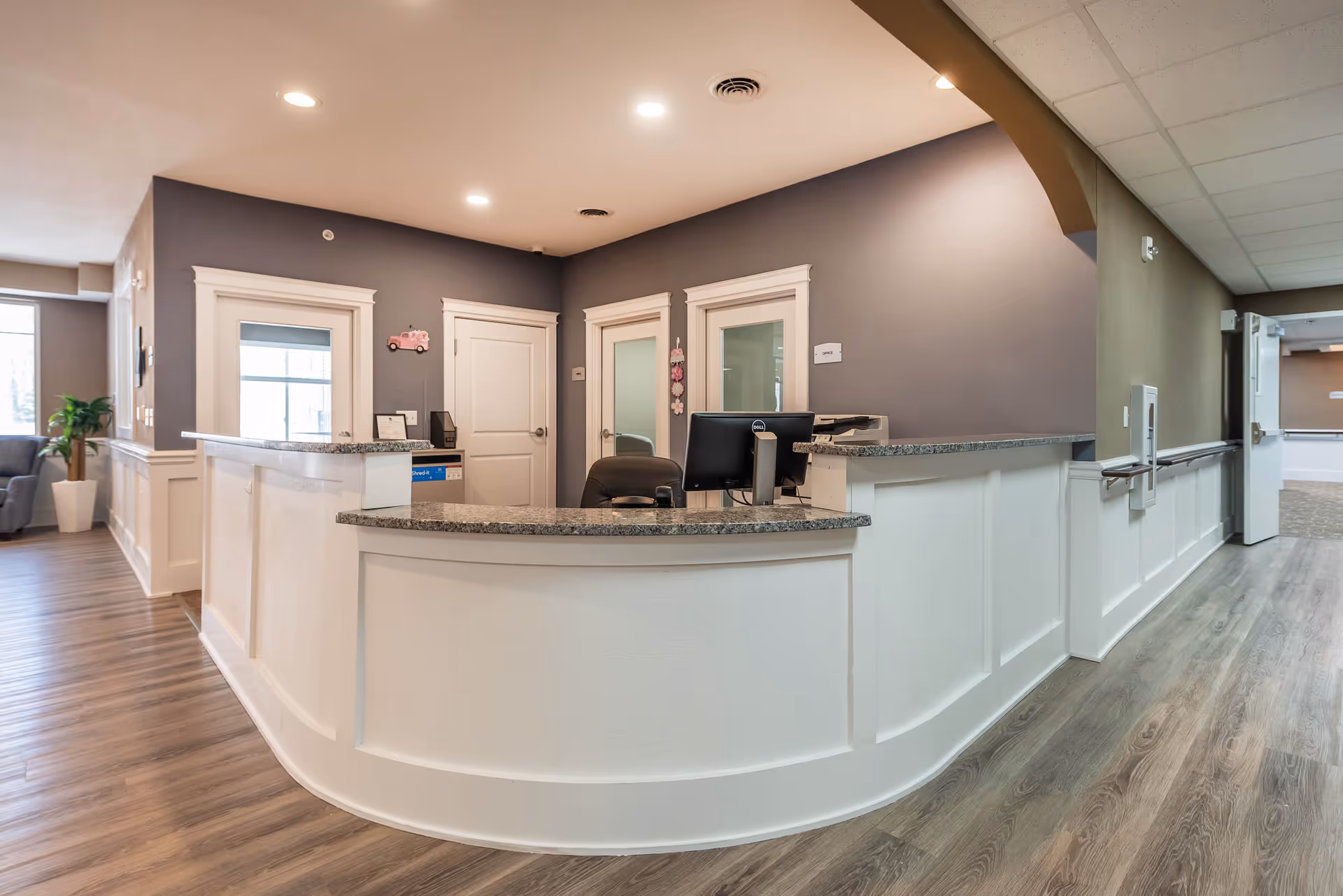 Reception desk area inside a senior living facility with a curved white counter, granite countertop, computer monitor, and office chair. The walls are painted gray with white trim around doors and windows. There is a hallway to the right and a seating area with a plant and armchair visible to the left.