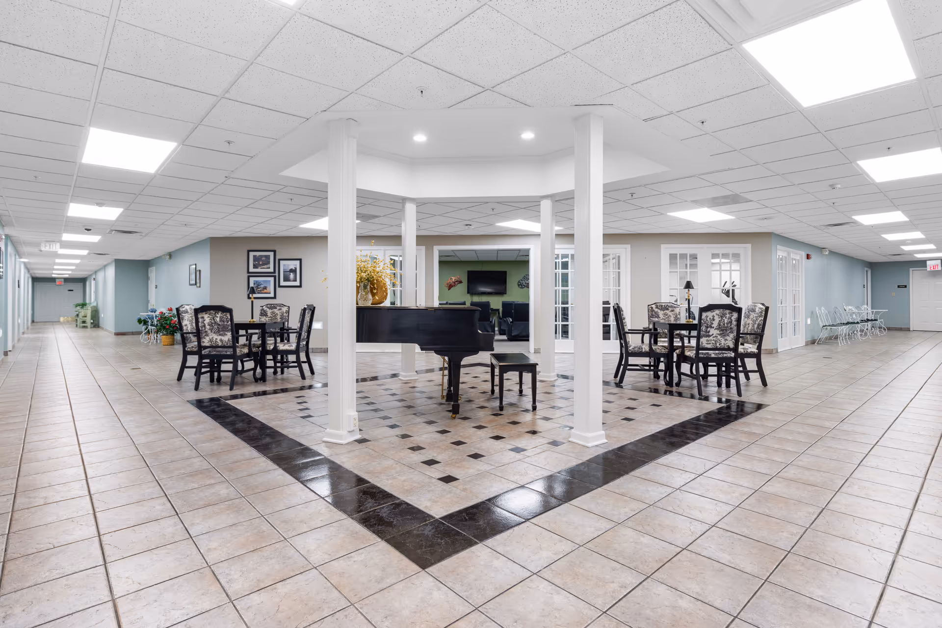 A spacious interior common area in an assisted living facility featuring a black grand piano in the center surrounded by white pillars. There are two seating areas with tables and chairs on either side of the piano. The floor is tiled with a decorative pattern around the piano. The walls are painted light blue and beige, and the ceiling has recessed lighting. In the background, there is a room with a television and additional seating visible through glass doors.