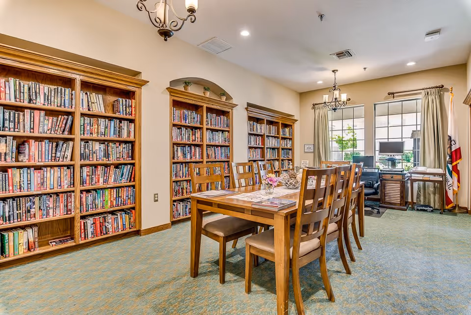 A well-lit library room with wooden bookshelves filled with books along the walls. In the center, there is a wooden table with six chairs around it, a small flower vase, and some magazines on the table. The room has carpeted flooring, two chandeliers hanging from the ceiling, and a window with curtains letting in natural light. A desk with a computer and office chair is positioned near the window, and a California state flag is visible in the corner.