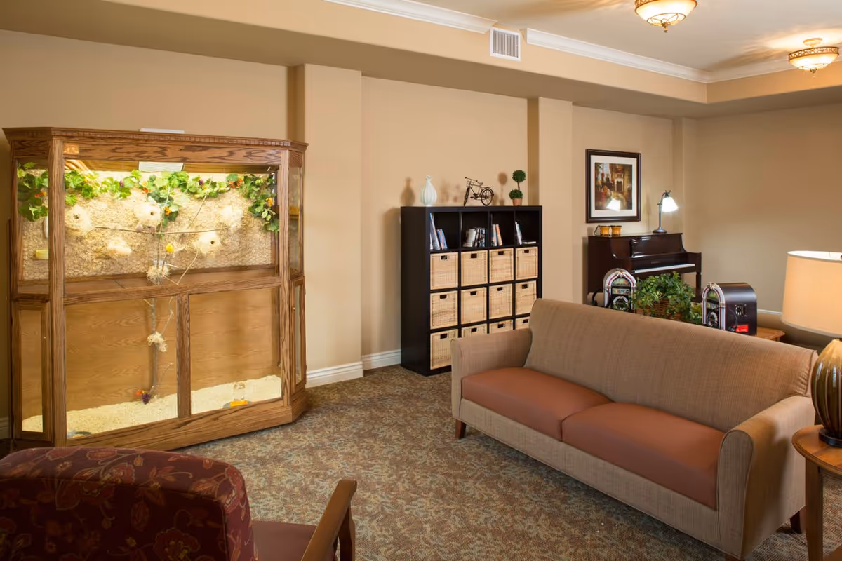 A cozy living room area with a beige couch, a patterned armchair, a wooden cabinet with multiple wicker baskets, a piano with a framed picture above it, and a large wooden terrarium with plants and nesting materials inside. The room has beige walls, carpeted floor, and ceiling lights.
