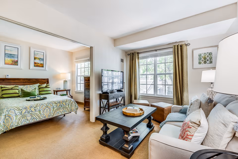 A bright and cozy senior living apartment featuring a living room and a bedroom. The living room has a beige sofa with multiple cushions, a dark wooden coffee table with books and a tray, a flat-screen TV on a stand, and large windows with green curtains letting in natural light. The adjacent bedroom has a wooden bed with a patterned bedspread and several pillows, two framed paintings on the wall, a bedside table with a lamp, and a window with blinds.