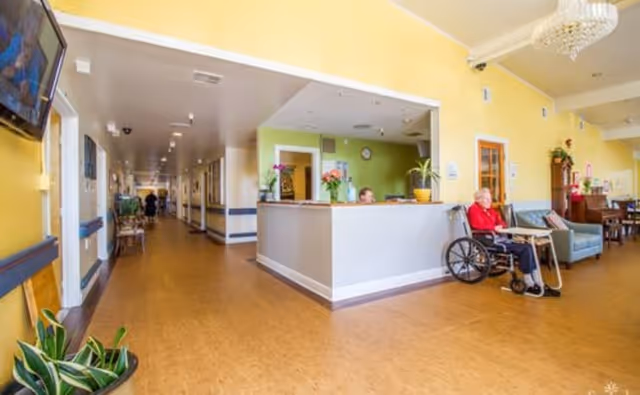 Interior view of a senior living facility reception area with a front desk, a person sitting behind the desk, and an elderly person in a wheelchair nearby. The hallway extends to the left with doors and a few people visible in the distance. The walls are painted yellow and green, and there are plants and chairs arranged around the space.