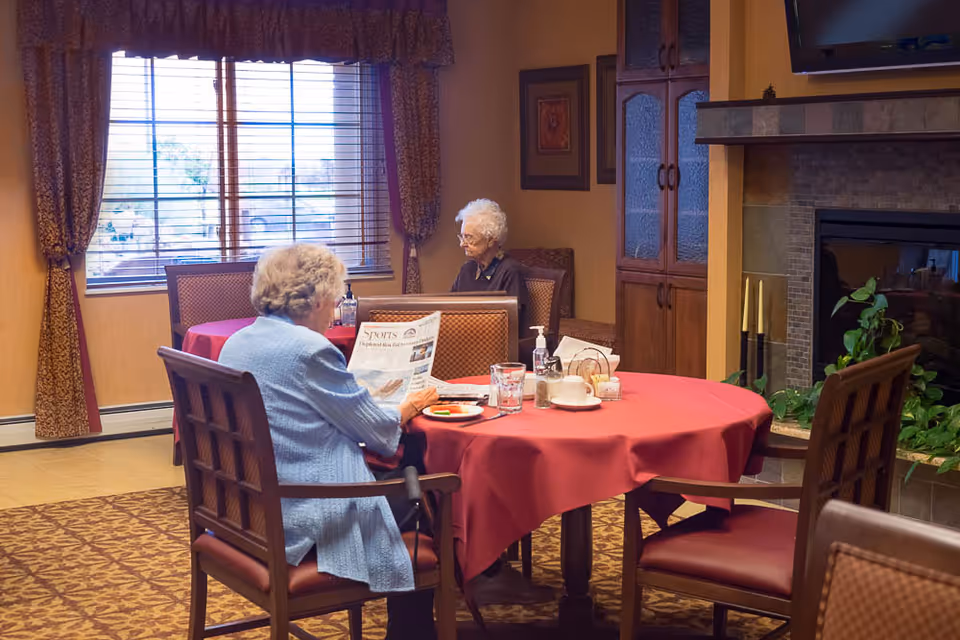 Two elderly women sitting at separate tables in a cozy dining room with red tablecloths, one woman reading a newspaper and the other looking ahead. The room features a large window with curtains, a fireplace, and wooden furniture.