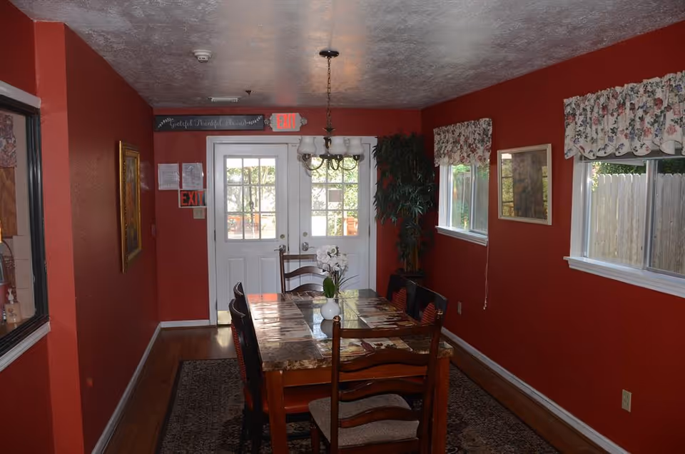 Dining room with a long wooden table and chairs, red walls, floral valances, a chandelier, and double glass doors.