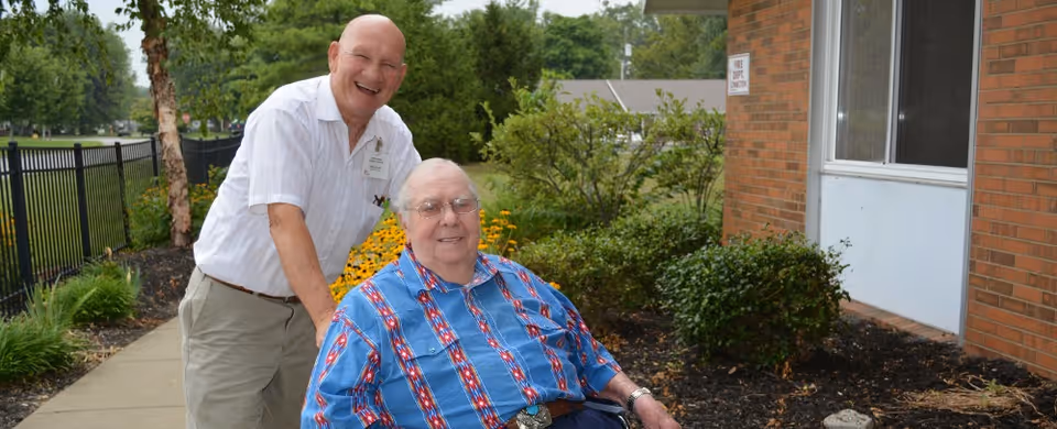 An elderly man in a wheelchair wearing a blue patterned shirt is outdoors near a brick building, accompanied by a smiling man standing behind him pushing the wheelchair. There are green bushes, flowers, and a black metal fence along a sidewalk in the background.