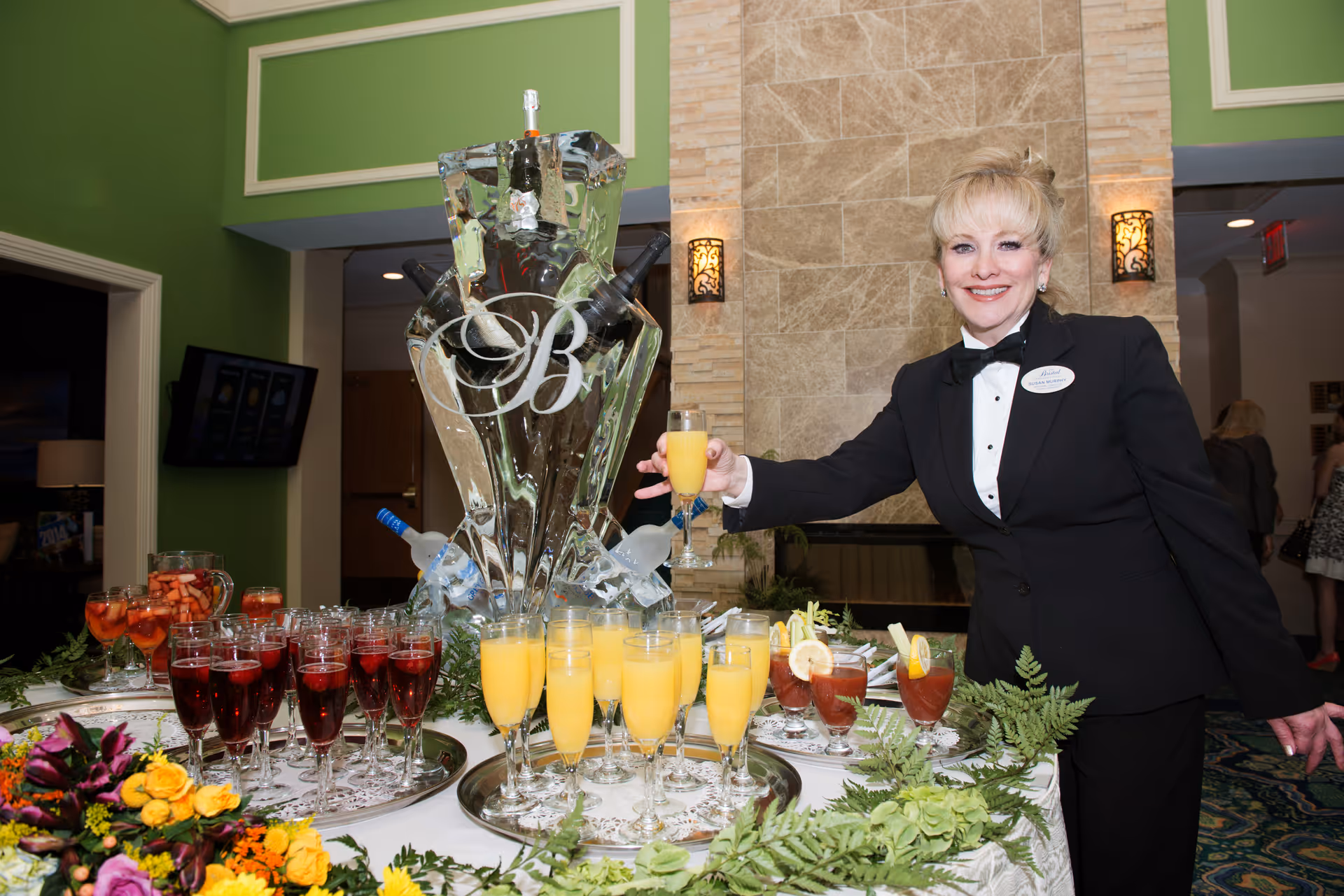 A woman dressed in a black tuxedo with a name tag is standing behind a table with various drinks including orange juice, red beverages, and iced tea garnished with lemon. The table is decorated with flowers and greenery, and there is a large ice sculpture with the letter 'B' and bottles of alcohol embedded in it. The setting appears to be an indoor event space with green walls and a stone fireplace in the background.