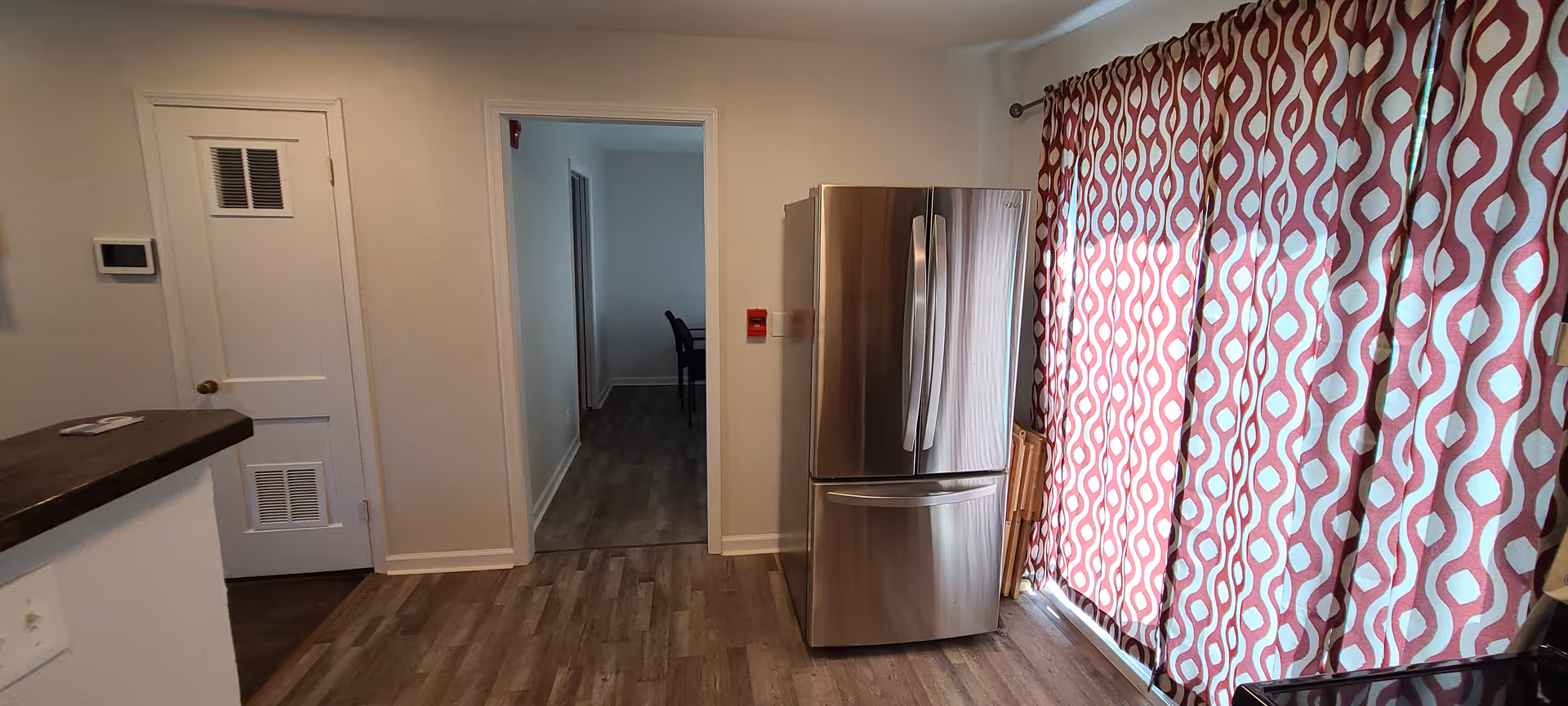 Interior view of a room with wood flooring, a stainless steel refrigerator, a door on the left wall, and a large window covered with red and white patterned curtains. A hallway is visible through an open doorway, leading to another room with a chair.