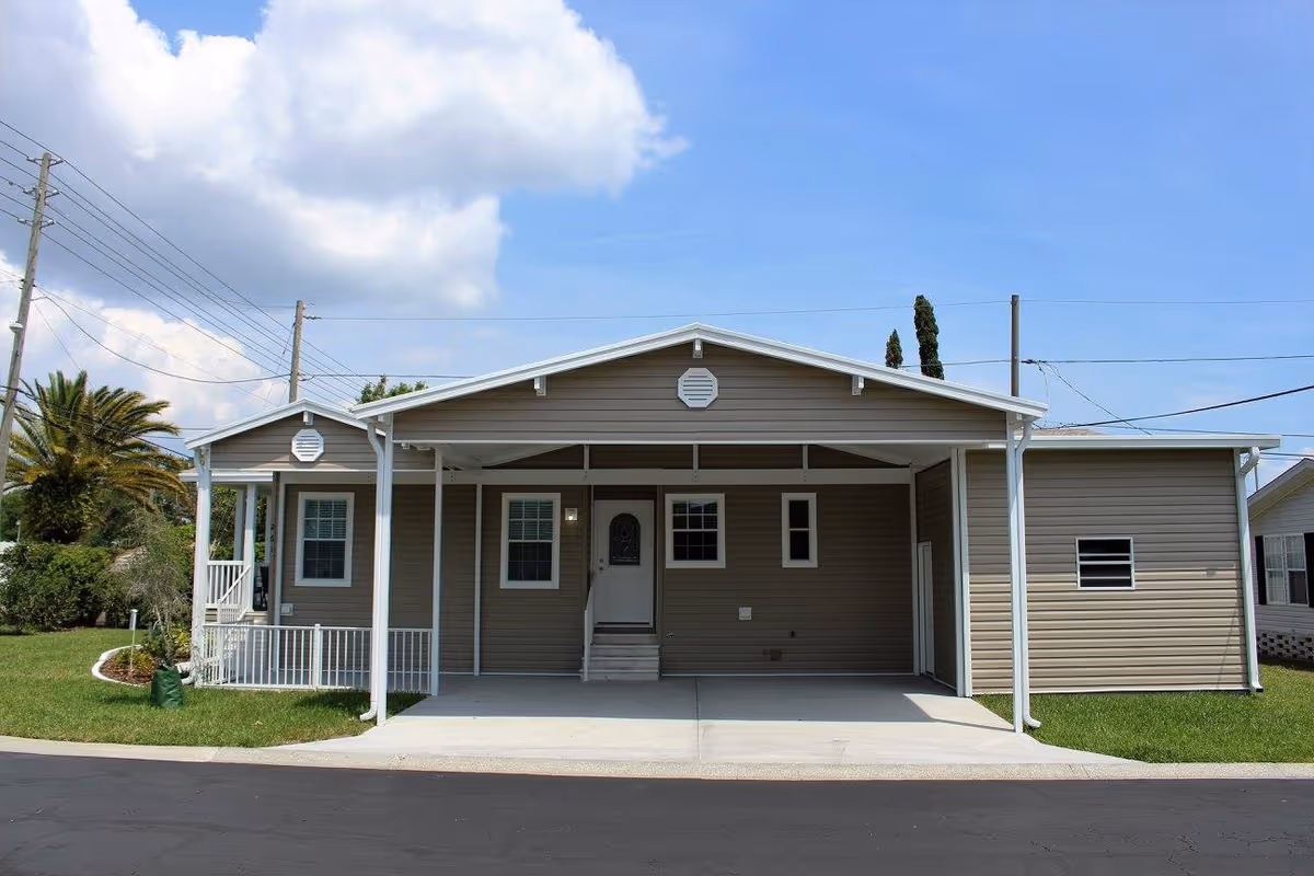Exterior view of a single-story manufactured home with beige siding and white trim. The home features a covered carport with a concrete driveway, several windows, a front door with steps, and a small porch area on the left side. The sky is partly cloudy and there is green grass surrounding the home.