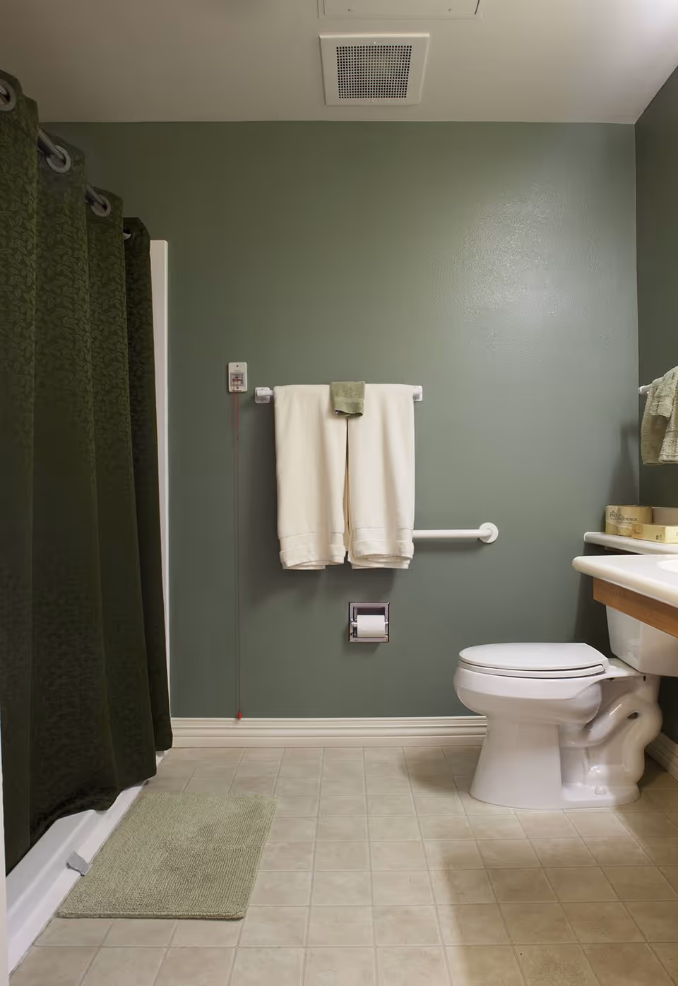 A clean bathroom with green walls and beige tiled floor. There is a white toilet on the right side next to a sink with a wooden cabinet. A white towel and a green washcloth hang on a towel rack above a grab bar. A green shower curtain is on the left side with a matching green bath mat on the floor. A pull cord emergency alert hangs next to the towel rack.