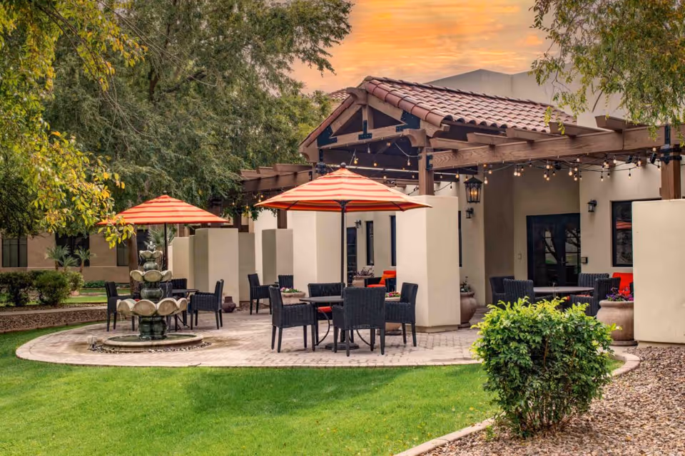 Outdoor courtyard with wicker tables and striped umbrellas centered around a tiered fountain beside a tiled-roof pergola.
