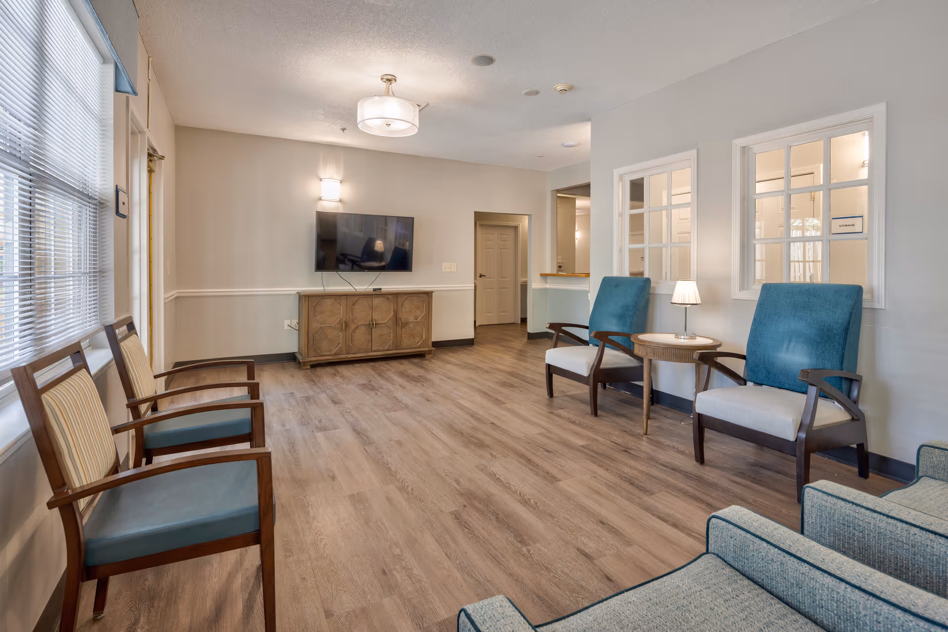 A bright and spacious living room area in TerraBella Hillsborough featuring wooden flooring, a wall-mounted flat-screen TV above a wooden cabinet, several armchairs with blue and beige cushions, a small round table with a lamp, and large windows with blinds allowing natural light to fill the room.