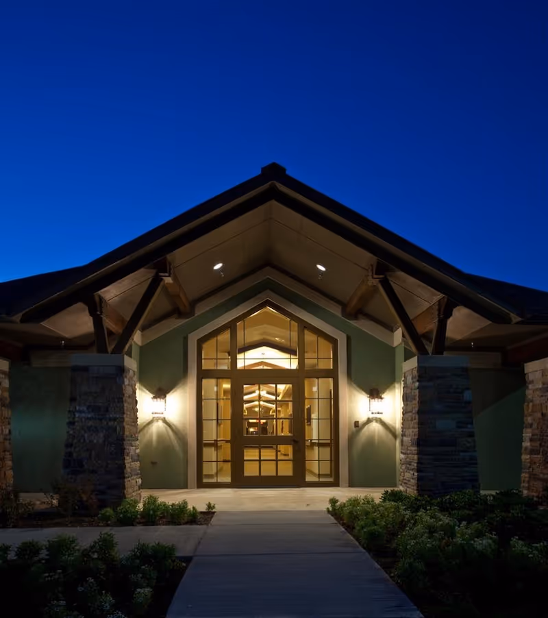 Illuminated front entrance with a peaked roof, stone columns and a lit glass doorway framed by landscaping under a deep blue evening sky.