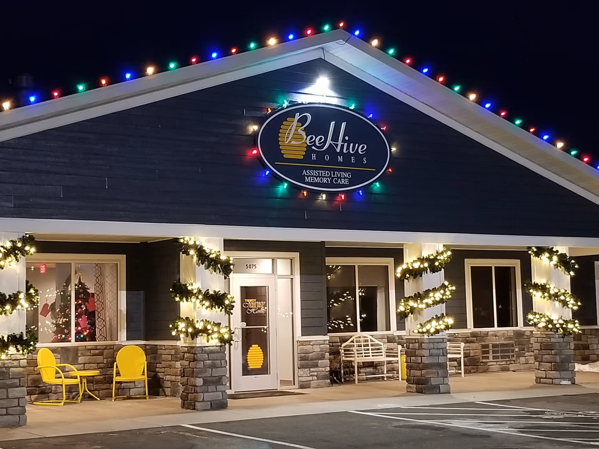 Front entrance of a BeeHive Homes assisted living building at night decorated with multicolored roof lights, illuminated garlands, and yellow patio chairs.