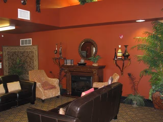 A cozy living room with dark brown leather sofas and a patterned armchair arranged around a wooden electric fireplace. The walls are painted a warm reddish-orange color and decorated with a round mirror above the fireplace, small shelves holding vases, and a large potted plant in the corner. The floor is carpeted with a patterned design.