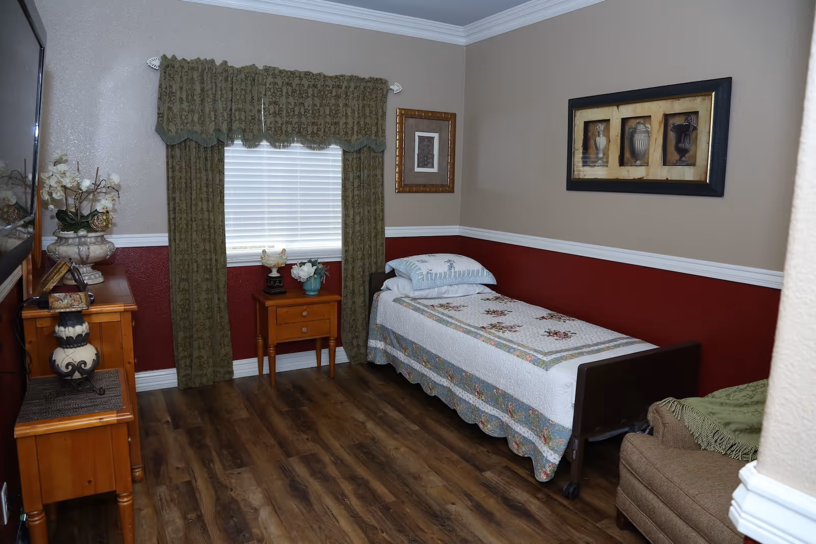 A small, neatly arranged bedroom with a single bed covered in a floral quilt. The room has wooden flooring, a window with green patterned curtains, a wooden nightstand with decorative items, a wooden dresser with a lamp and flowers, and framed artwork on the walls.