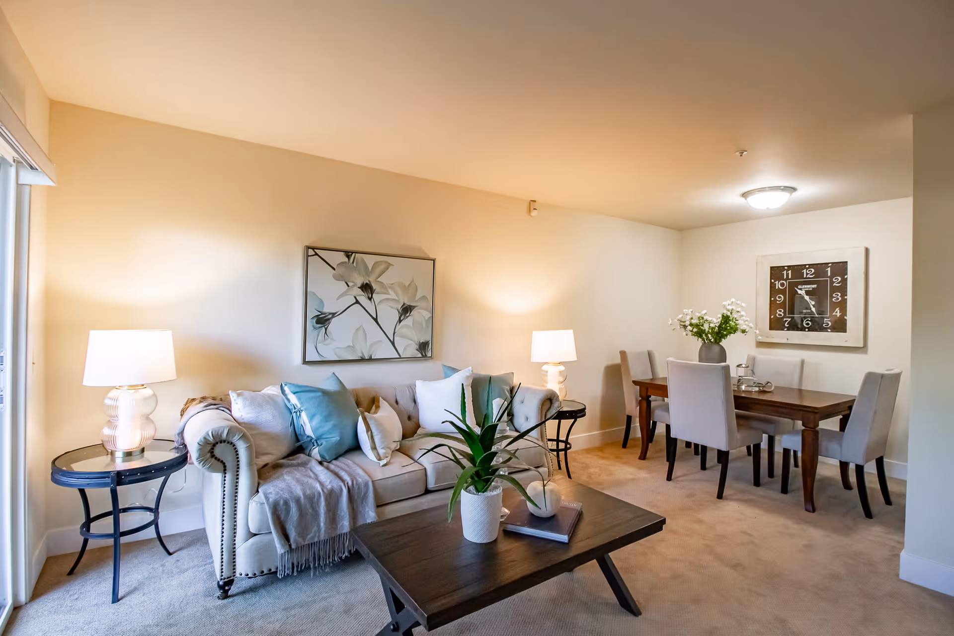 A cozy living and dining area featuring a beige tufted sofa with multiple cushions and a throw blanket, two side tables with lamps, a wooden coffee table with a potted plant and books, and a dining table with six upholstered chairs. The walls are light-colored with a floral painting above the sofa and a large clock above the dining table.